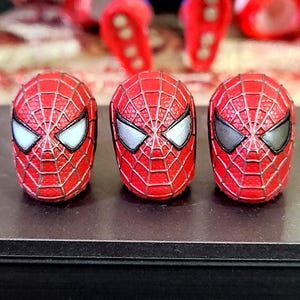 Puede incluir: Tres cabezas de figura de acción rojas con una superficie texturizada y un patrón de telaraña plateado en relieve. Cada cabeza tiene lentes oculares distintas: la izquierda tiene lentes blancas, la central plateadas y la derecha gris oscuro.