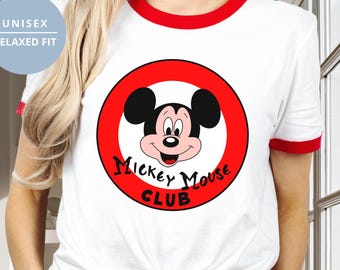 Puede incluir: Una camiseta blanca con ribetes rojos en el cuello y las mangas. El centro presenta un gráfico circular rojo con un contorno negro que contiene una ilustración de Mickey Mouse y el texto negro Mickey Mouse Club. La camiseta tiene un corte holgado.