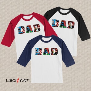 Op de afbeelding: Drie baseball shirts met het woord "DAD" gespeld in stripfiguren. De shirts zijn wit met rode, donkerblauwe en zwarte mouwen.