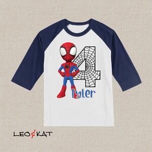 May include: A white and navy blue baseball t-shirt with a cartoon image of Spiderman standing in front of a large number 4 with a spiderweb design. The name Tyler is written below the number.