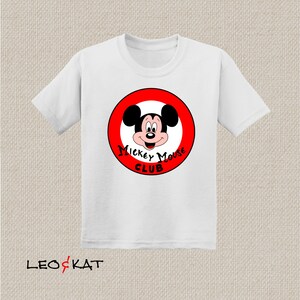 以下が含まれることがあります： 赤い円の中にミッキーマウスの顔が描かれた白いTシャツ。円には「Mickey Mouse Club」という文字が書かれています。