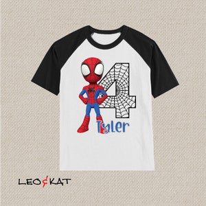 May include: White and black raglan t-shirt with a cartoon image of Spiderman in a red and blue suit standing in front of a black and white number 4 with a spider web design. The text "Tyler" is printed below the number.