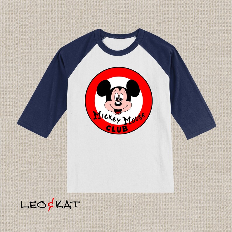 以下が含まれることがあります： 白と紺のベースボールTシャツ。赤い円の中にミッキーマウスと「Mickey Mouse Club」の文字がデザインされています。