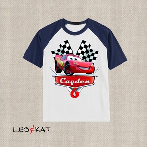 Może przedstawiać: Biało-granatowa koszulka typu raglan z czerwonym samochodem wyścigowym, szachownicą i nadrukiem imienia "Cayden" z przodu.