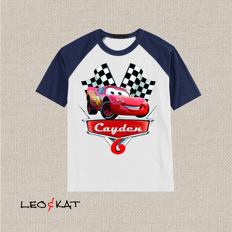 Może przedstawiać: Biało-granatowa koszulka typu raglan z czerwonym samochodem wyścigowym, szachownicą i nadrukiem imienia "Cayden" z przodu.