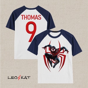 May include: White and navy blue raglan t-shirt with a red number 9 and the name "THOMAS" on the back. The front of the shirt features a red and black illustration of Spider-Man.