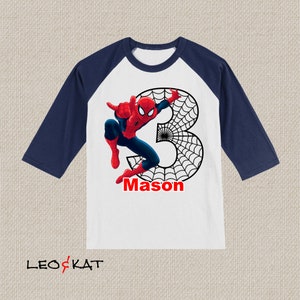 Puede incluir: Una camiseta de b&eacute;isbol blanca y azul marino con un gr&aacute;fico de Spiderman rojo y azul, una telara&ntilde;a negra y el n&uacute;mero 3. La camiseta tambi&eacute;n tiene el nombre "Mason" impreso en ella.