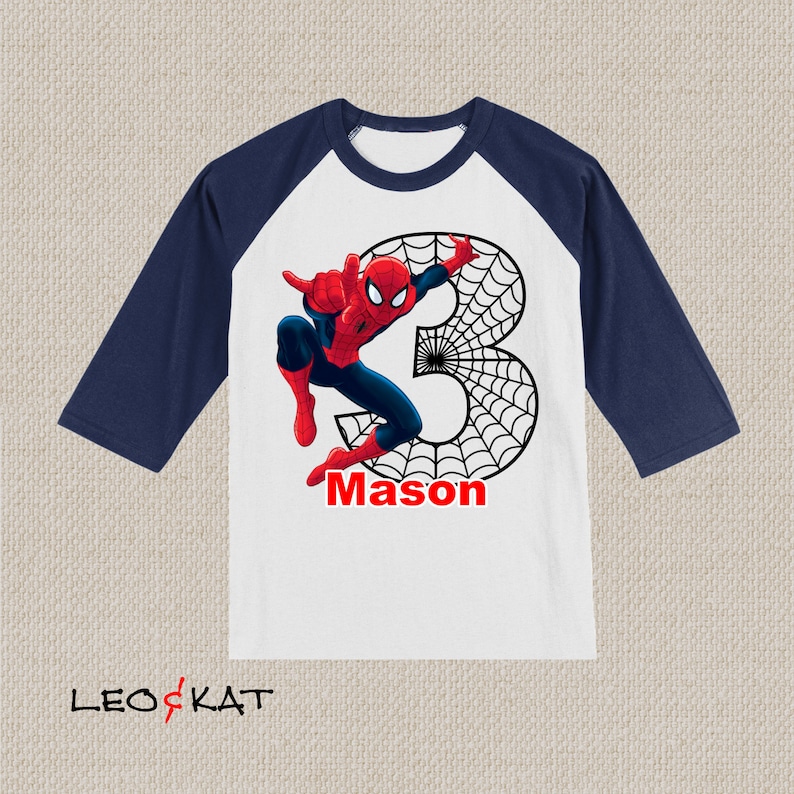 Puede incluir: Una camiseta de b&eacute;isbol blanca y azul marino con un gr&aacute;fico de Spiderman rojo y azul, una telara&ntilde;a negra y el n&uacute;mero 3. La camiseta tambi&eacute;n tiene el nombre "Mason" impreso en ella.