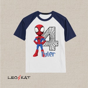 May include: A white and navy blue raglan t-shirt with a cartoon image of Spiderman in a red and blue suit. The number 4 is behind Spiderman with a black spiderweb design. The name Tyler is written below Spiderman.