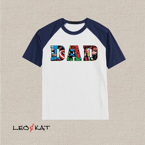 Op de afbeelding: Wit T-shirt met donkerblauwe raglanmouwen. Het shirt heeft een afbeelding van het woord "DAD" gemaakt van stripboekafbeeldingen van superhelden.