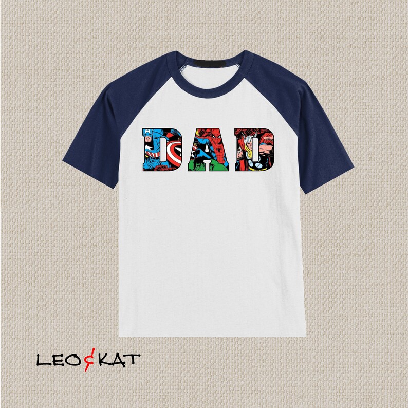 Op de afbeelding: Wit T-shirt met donkerblauwe raglanmouwen. Het shirt heeft een afbeelding van het woord "DAD" gemaakt van stripboekafbeeldingen van superhelden.