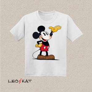 以下が含まれることがあります： ミッキーマウスの漫画のキャラクターのイラストが描かれた白いTシャツ。ミッキーは、白いボタンが付いた特徴的な赤いショートパンツ、黄色の手袋、茶色の靴を履いています。シャツの下部には「LEO & KAT」の文字があります。