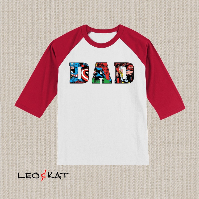 Op de afbeelding: Wit en rood baseballshirt met het woord "DAD" in grote letters. De letters zijn gemaakt van stripboekafbeeldingen van superhelden.