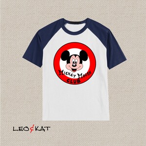 以下が含まれることがあります： 白と紺色のラグランスリーブTシャツ。赤い円形のグラフィックにはミッキーマウスと「ミッキーマウスクラブ」の文字が描かれています。