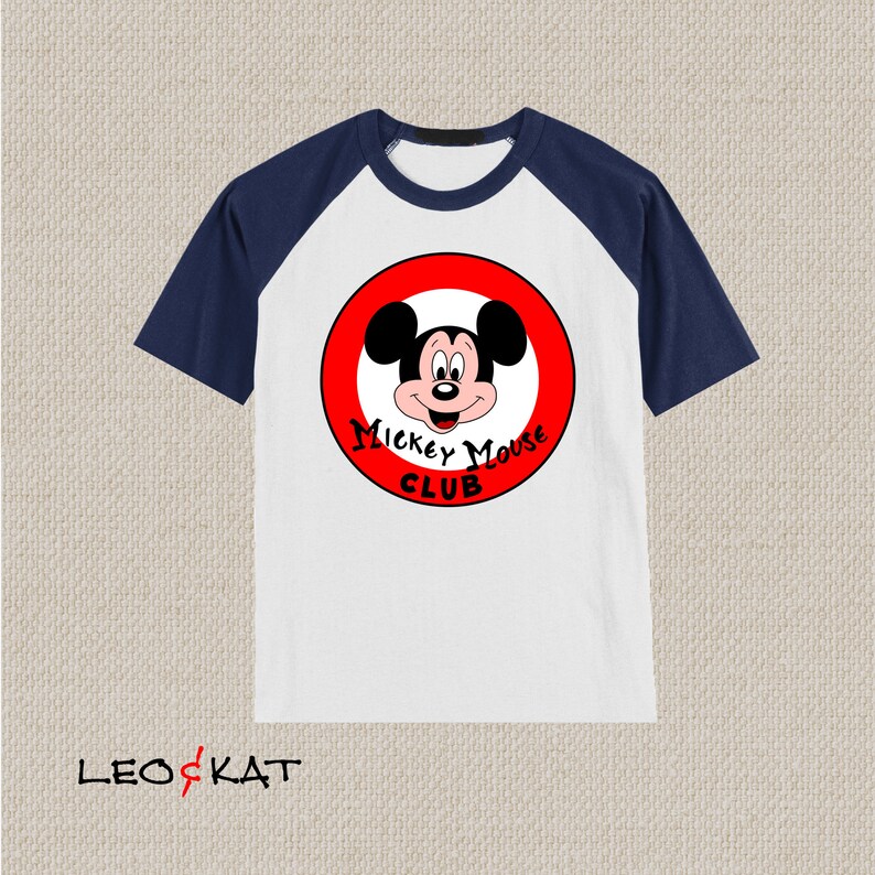以下が含まれることがあります： 白と紺色のラグランスリーブTシャツ。赤い円形のグラフィックにはミッキーマウスと「ミッキーマウスクラブ」の文字が描かれています。