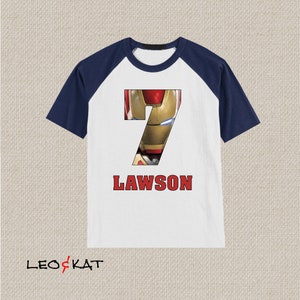 Pu&ograve; includere: Maglietta a maniche raglan bianca e blu navy con un numero 7 rosso che presenta un'immagine del volto di Iron Man. Il testo "LAWSON" &egrave; stampato in rosso sotto il numero.