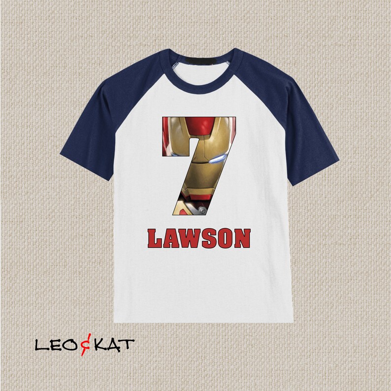 Pu&ograve; includere: Maglietta a maniche raglan bianca e blu navy con un numero 7 rosso che presenta un'immagine del volto di Iron Man. Il testo "LAWSON" &egrave; stampato in rosso sotto il numero.