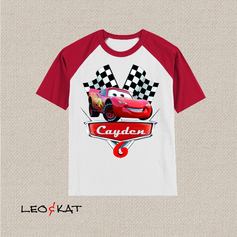 Może przedstawiać: Czerwono-biała koszulka typu raglan z nadrukiem kresk&oacute;wkowego wizerunku Złomka McQueen z filmu Auta, szachownicy i imieniem "Cayden" na przodzie.