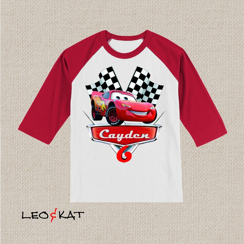 Może przedstawiać: Czerwono-biała koszulka bejsbolowa z czerwonym samochodem wyścigowym i szachownicą. Koszulka ma napis "Cayden" i numer "6".