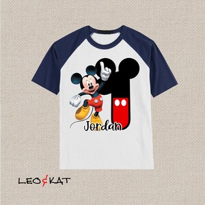 Peut inclure: T-shirt &agrave; manches raglan blanche et bleu marine avec un motif Mickey Mouse et le nom "Jordan" imprim&eacute; sur le devant.