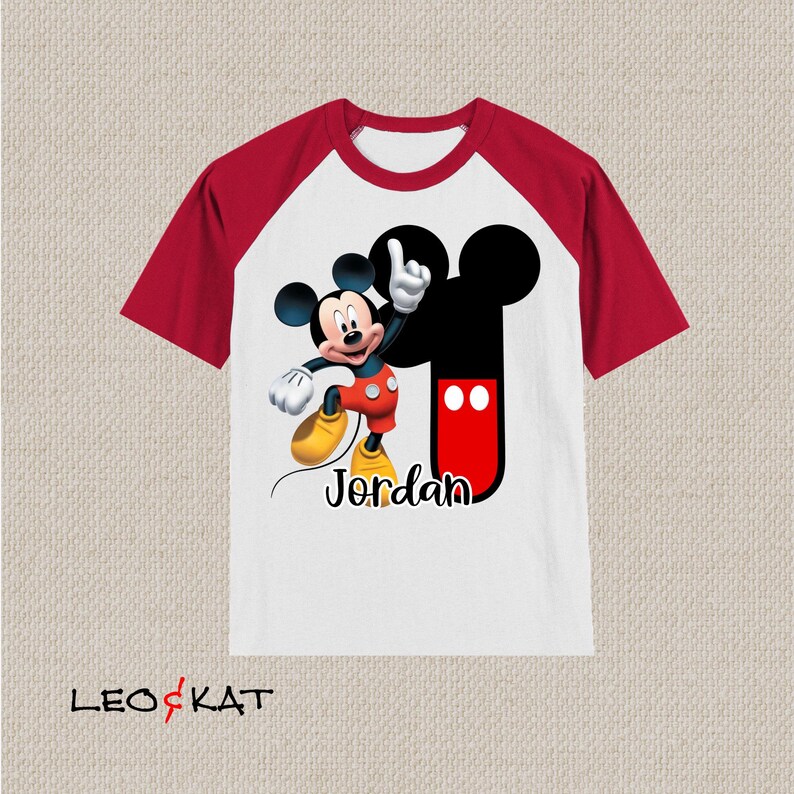 Peut inclure: T-shirt &agrave; manches raglan blanche et rouge avec un motif Mickey Mouse noir et rouge. Le t-shirt porte le nom "Jordan" imprim&eacute; sur le devant.