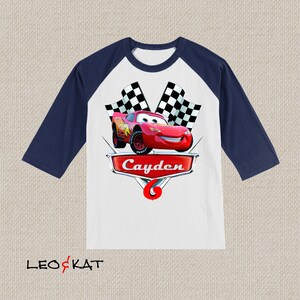 Może przedstawiać: Biało-granatowa koszulka bejsbolowa z czerwonym samochodem wyścigowym i szachownicą. Na koszulce nadrukowano imię "Cayden" i numer "6".