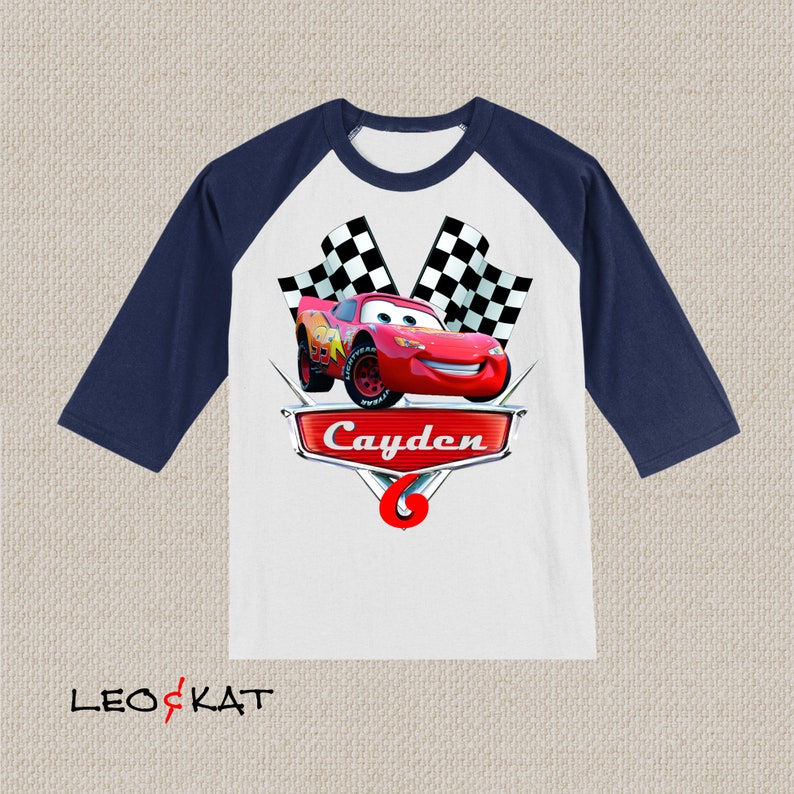 Może przedstawiać: Biało-granatowa koszulka bejsbolowa z czerwonym samochodem wyścigowym i szachownicą. Na koszulce nadrukowano imię "Cayden" i numer "6".