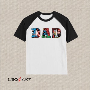 Op de afbeelding: Wit en zwart baseballshirt met een afbeelding van het woord "DAD" gemaakt van stripfiguren.