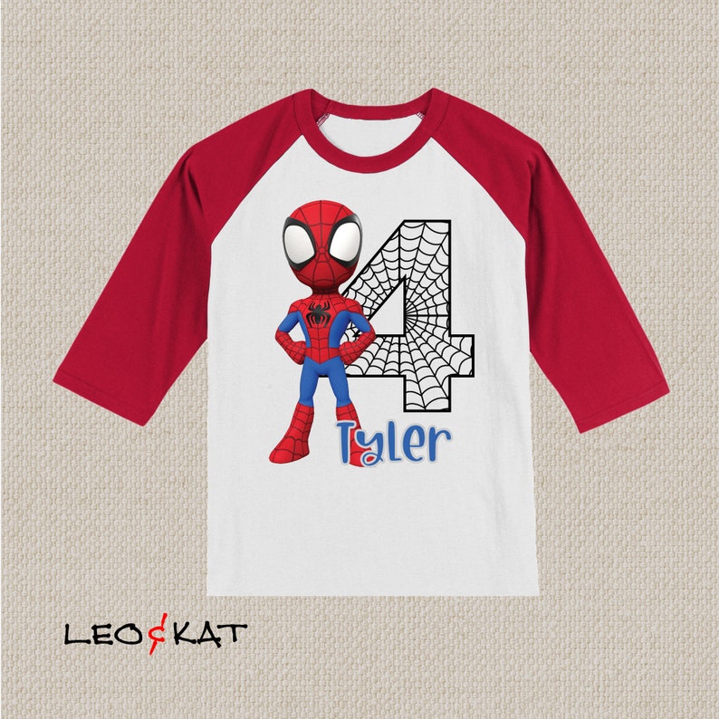 May include: A white and red baseball tee shirt with a red and blue Spiderman character and a black number 4 with a spiderweb design. The name Tyler is written in blue below the number.