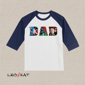 Op de afbeelding: Een wit en donkerblauw baseballshirt met het woord "DAD" erop gedrukt. De letters zijn gemaakt van afbeeldingen van superhelden uit Marvel Comics.