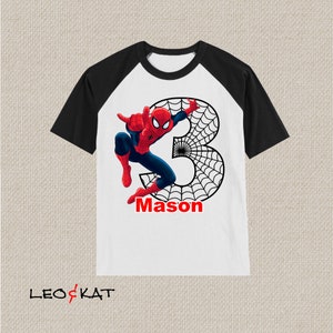 Puede incluir: Una camiseta blanca y negra de manga raglan con un gr&aacute;fico de Spiderman rojo y azul y el n&uacute;mero 3. El gr&aacute;fico tambi&eacute;n incluye una telara&ntilde;a negra. El nombre "Mason" est&aacute; escrito en rojo debajo del gr&aacute;fico.