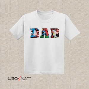 Op de afbeelding: Wit T-shirt met het woord "DAD" gespeld in kleurrijke stripfiguren. De figuren zijn van Marvel Comics, waaronder Captain America, Spider-Man en Thor.