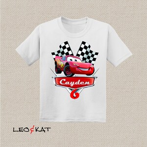 Może przedstawiać: Biała koszulka z czerwonym samochodem wyścigowym, szachownicą i imieniem "Cayden" w kolorze czerwonym.