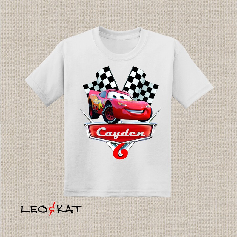 Może przedstawiać: Biała koszulka z czerwonym samochodem wyścigowym, szachownicą i imieniem "Cayden" w kolorze czerwonym.