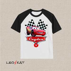 Może przedstawiać: Biało-czarna koszulka bejsbolowa z czerwonym samochodem wyścigowym i szachownicą. Samoch&oacute;d ma uśmiechniętą twarz, a na koszulce widnieje napis "Cayden".
