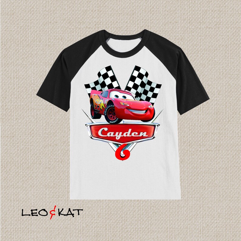 Może przedstawiać: Biało-czarna koszulka bejsbolowa z czerwonym samochodem wyścigowym i szachownicą. Samoch&oacute;d ma uśmiechniętą twarz, a na koszulce widnieje napis "Cayden".