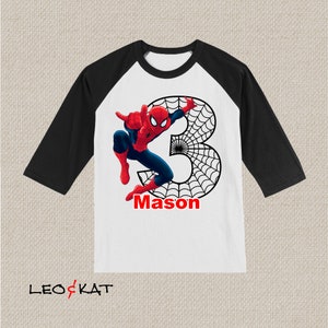 Puede incluir: Una camiseta de b&eacute;isbol blanca y negra con un gr&aacute;fico de Spiderman rojo y azul. El n&uacute;mero 3 est&aacute; en el fondo con un dise&ntilde;o de telara&ntilde;a negra. El nombre Mason est&aacute; escrito en rojo debajo del gr&aacute;fico.