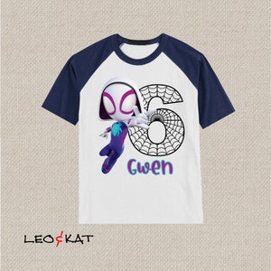 May include: White and navy blue raglan t-shirt featuring a cartoon illustration of Spider-Gwen with the number 6 and the name "Gwen". The graphic is in vibrant colours, with a spider web design incorporated into the number.