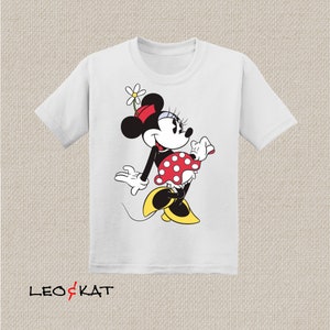 K&ouml;nnte beinhalten: Wei&szlig;es T-Shirt mit einem Cartoon-Bild von Minnie Mouse, die ein rot-wei&szlig; gepunktetes Kleid und eine rote Schleife tr&auml;gt. Sie h&auml;lt eine wei&szlig;e Blume in ihren Haaren und hat einen gelben Schuh an einem Fu&szlig;.