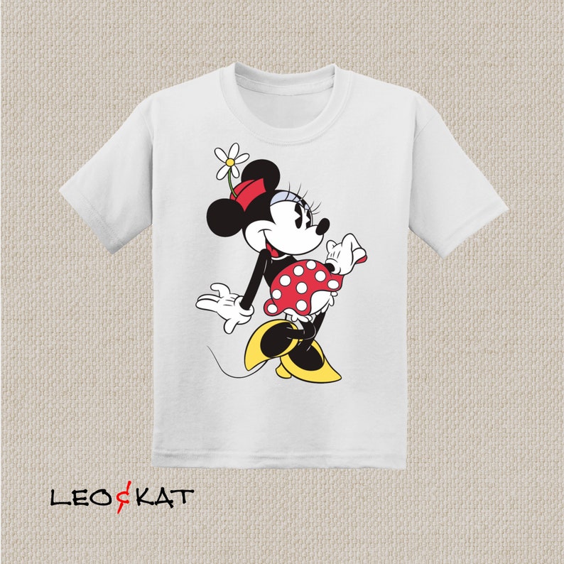 K&ouml;nnte beinhalten: Wei&szlig;es T-Shirt mit einem Cartoon-Bild von Minnie Mouse, die ein rot-wei&szlig; gepunktetes Kleid und eine rote Schleife tr&auml;gt. Sie h&auml;lt eine wei&szlig;e Blume in ihren Haaren und hat einen gelben Schuh an einem Fu&szlig;.