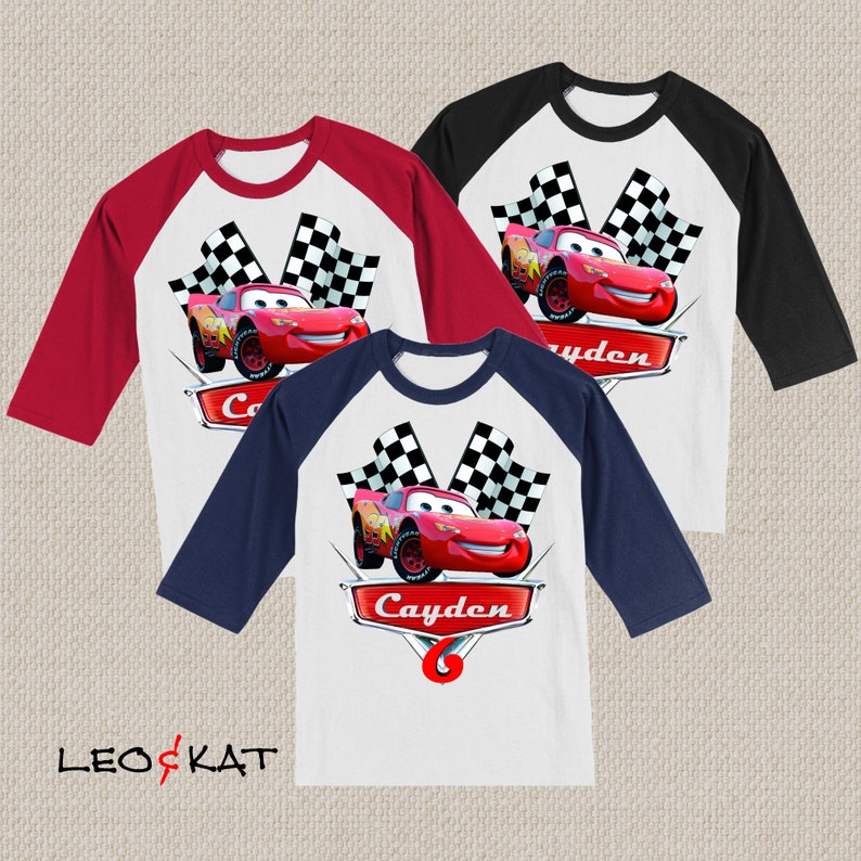 Może przedstawiać: Trzy białe, czerwone i niebieskie koszulki bejsbolowe z wzorem czerwonego samochodu wyścigowego i szachownicy. Koszulki mają nadrukowane imię "Cayden" i numer "6".