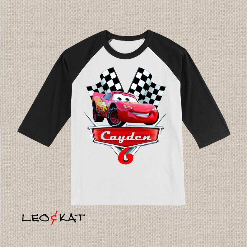 Może przedstawiać: Biało-czarna koszulka w stylu bejsbolowym z czerwonymi i czarnymi szachownicami, czerwonym samochodem wyścigowym i imieniem "Cayden" na srebrnym banerze.