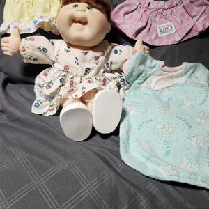 Puede incluir: Una muñeca de pelo castaño y ojos verdes con vestido de flores y zapatos blancos, acompañada de un top amarillo, una falda rosa con parche CPK y una manta verde claro con el texto It's a Girl e ilustraciones de bebés.