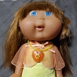 Könnte beinhalten: Eine Cabbage Patch Kids Puppe mit braunen Haaren, blauen Augen und einem rosa-gelben Outfit. Die Puppe trägt eine Halskette mit einem herzförmigen Anhänger.