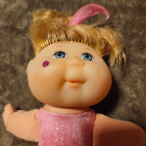 Peut inclure: Une poupée Cabbage Patch Kids aux cheveux blonds portant un justaucorps rose pailleté. La poupée a les yeux bleus et un autocollant de coccinelle sur la joue.