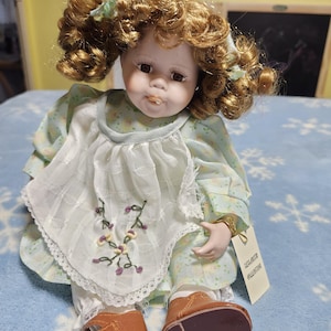 Puede incluir: Muñeca de porcelana con cabello castaño rizado y ojos marrones. La muñeca viste un vestido floral verde claro con un babero blanco adornado con flores bordadas. Lleva zapatos de cuero marrón. Una etiqueta está sujeta a la muñeca.