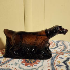 May include: A dark brown glass bottle shaped like a dog, with a black leather collar. The bottle is sitting on a patterned fabric.