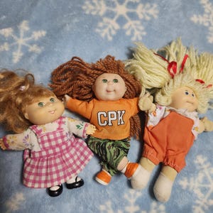 May include: Three Cabbage Patch Kids dolls. One doll wears a pink and white plaid dress, another wears an orange shirt with "CPK" on it and camouflage trousers, and the third doll wears an orange jumpsuit. All dolls have yarn hair.