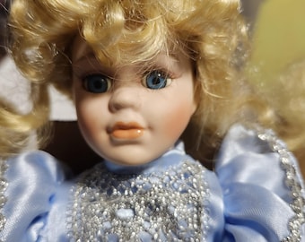May include: A porcelain doll with blonde, curly hair and blue eyes. The doll wears a light blue satin dress with silver lace detailing around the neckline and sleeves. The doll's face is delicately painted.
