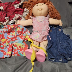 Pode incluir: Uma boneca Cabbage Patch Kids com cabelo de lã castanho-avermelhado, vestindo um vestido rosa com flores e sapatos rosa. Estão incluídos três conjuntos adicionais: um vestido vermelho com flores e renda branca, calções vermelhos com estampado de dinossauros e jardineiras de ganga azul.